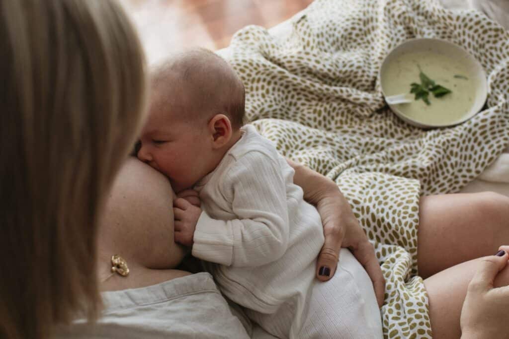 Village For Mama A person breastfeeding an infant while sitting on a bed with a bowl of soup on a nearby patterned blanket.