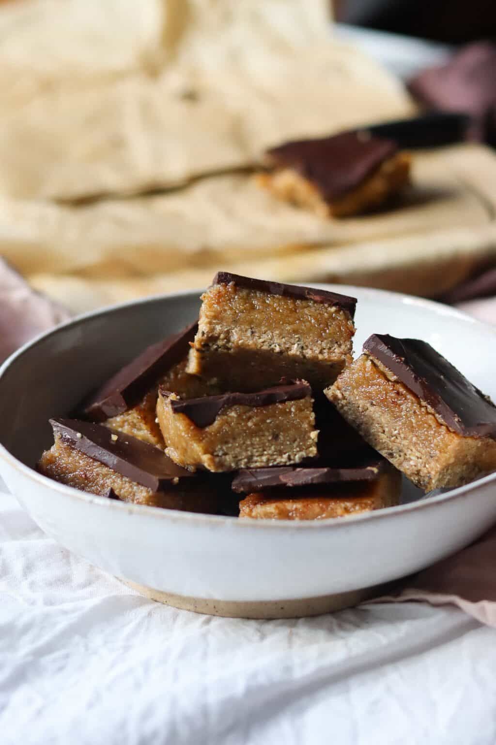 Village For Mama A white bowl filled with square-shaped layered dessert bars, featuring dark chocolate on top and a crumbly base, is set on a table. A blurred piece of parchment paper is in the background.
