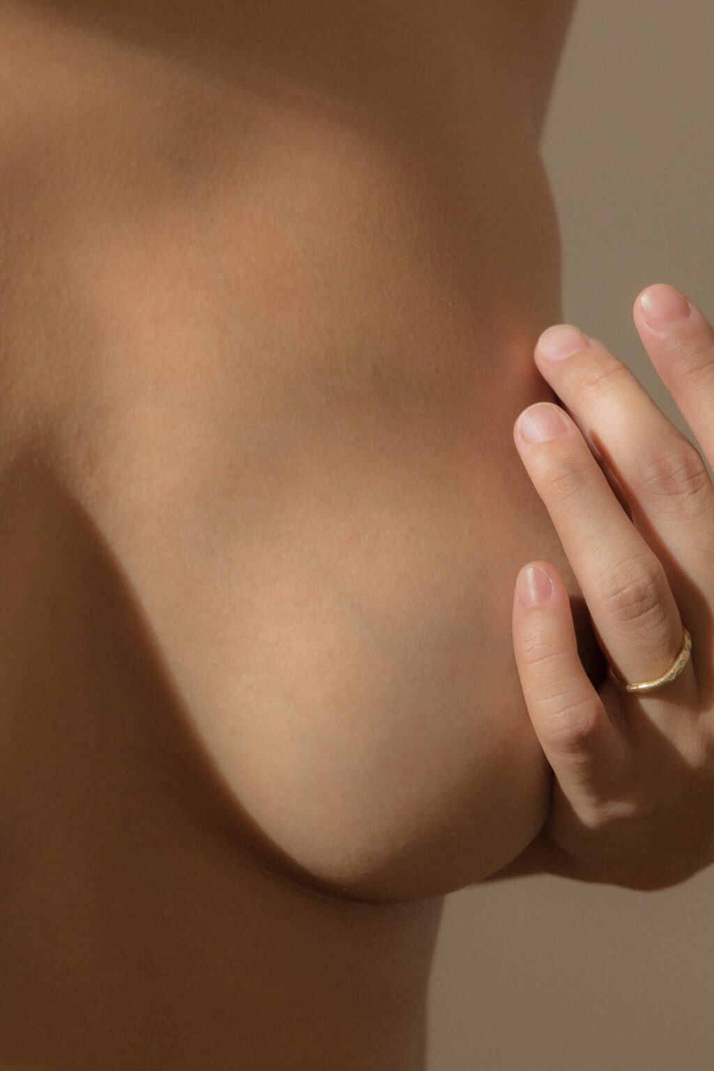 Village For Mama A close-up image of a person with their hand gently touching the side of their bare breast. The person is wearing a simple gold ring on their finger.