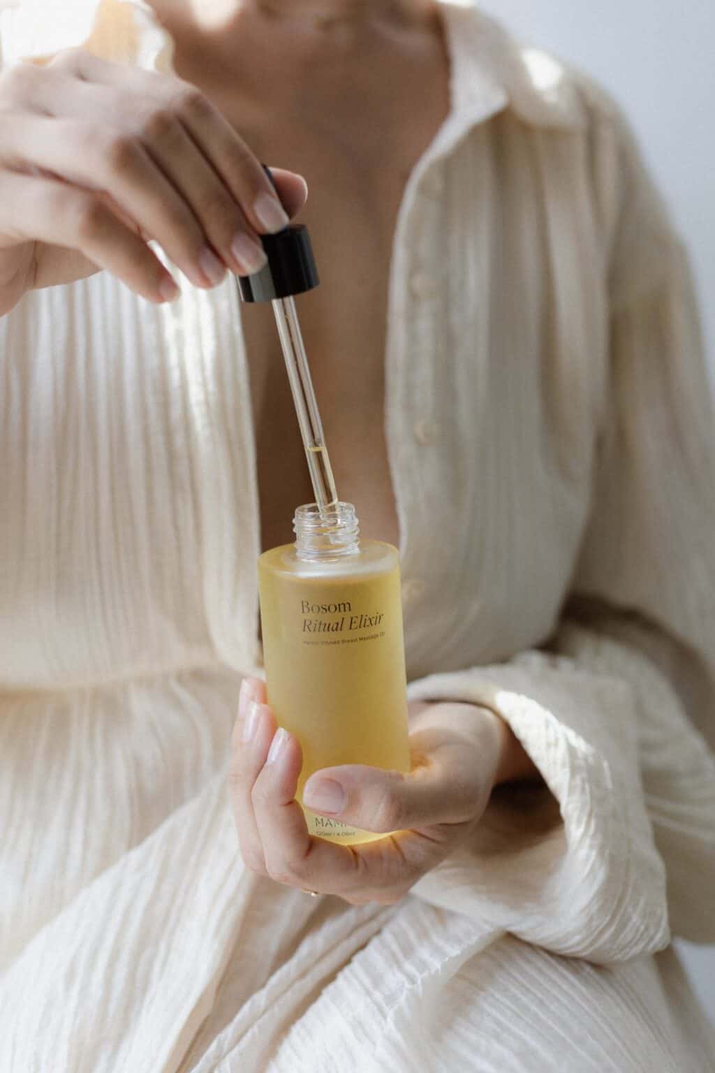 Village For Mama A person in a beige, textured robe holding a dropper above a bottle labeled "Bosom Ritual Elixir.
