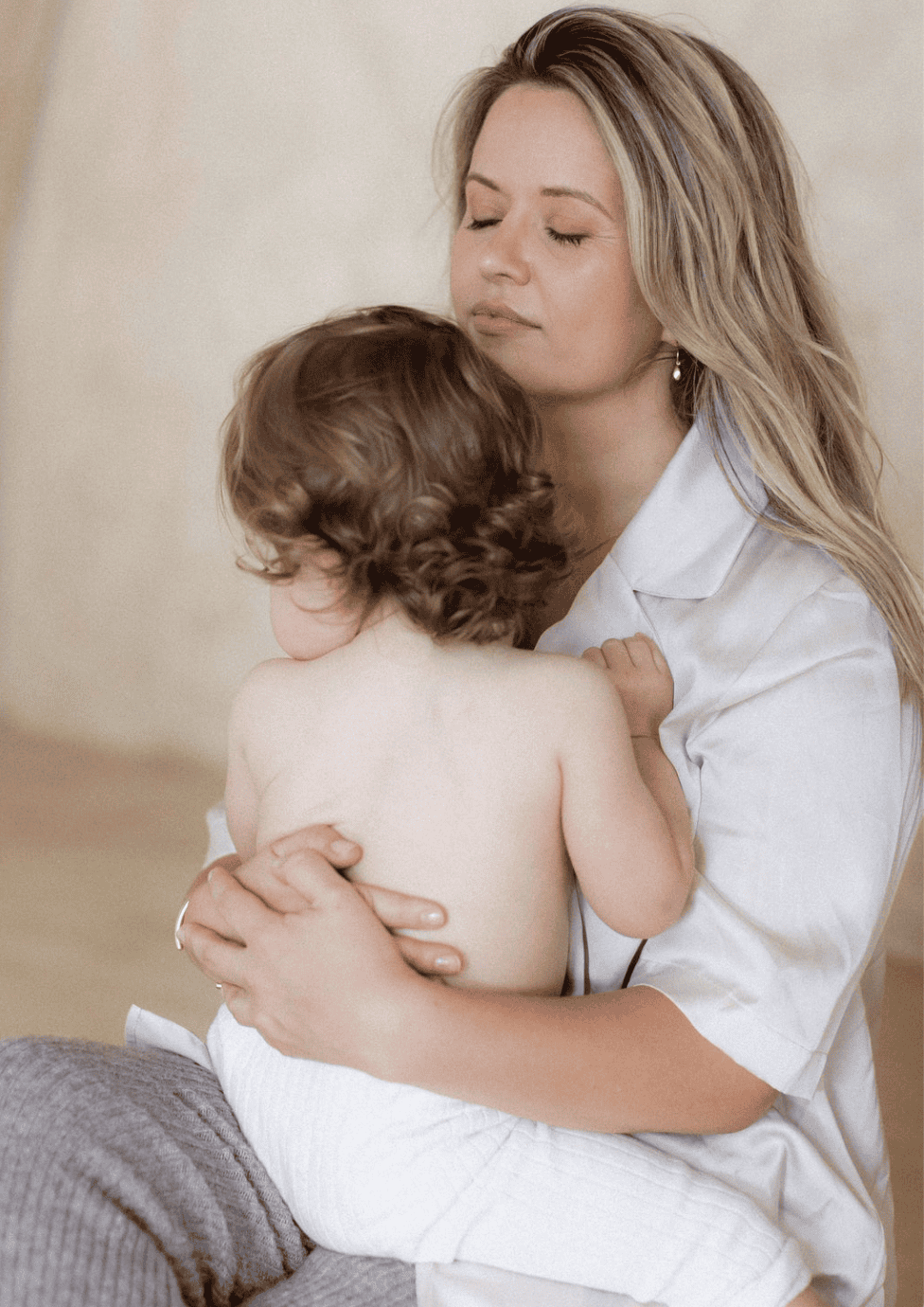 Village For Mama A woman with long blonde hair holds a toddler with curly hair on her lap. The toddler is shirtless while the woman wears a white shirt and gray pants. Both have calm expressions.