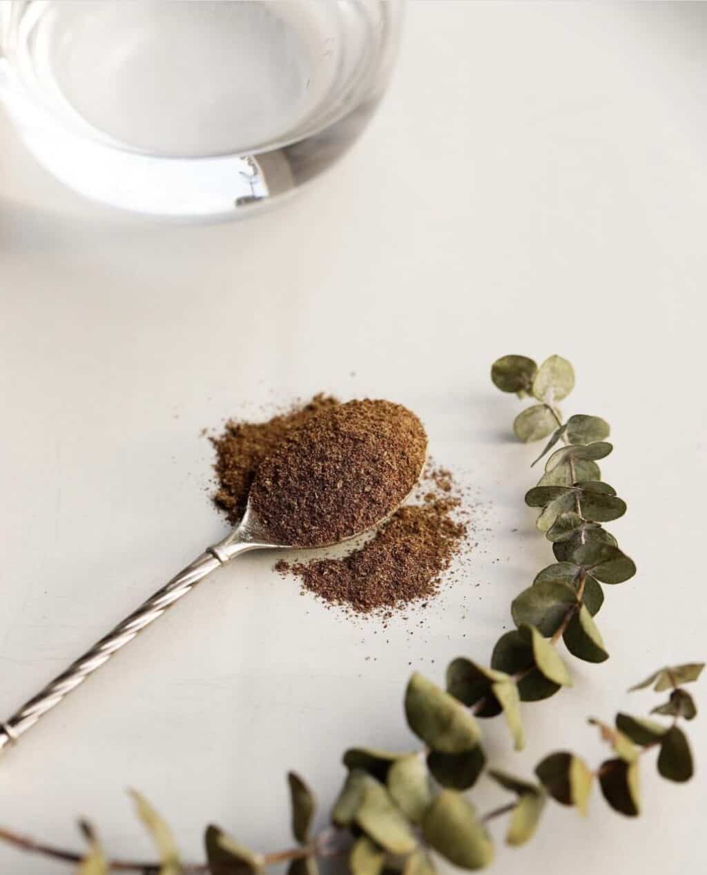 Village For Mama A spoon with a pile of ground spice on a white surface, accompanied by a glass of water and a sprig of green leaves.