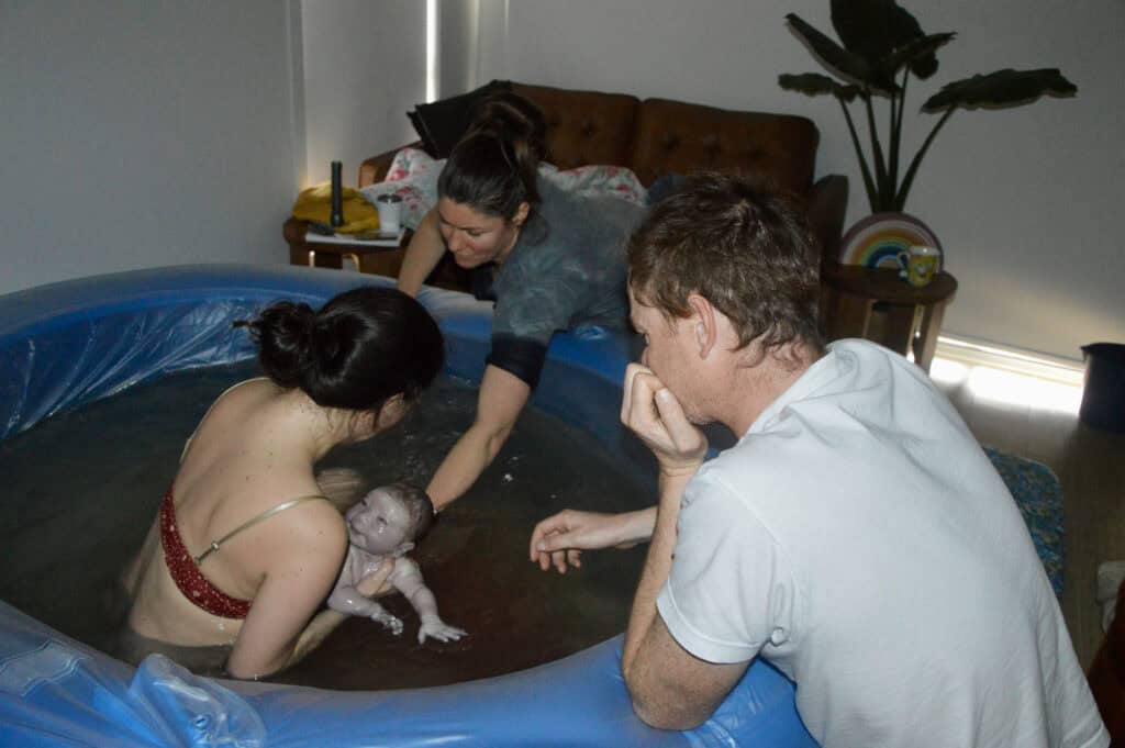 Village For Mama A woman in a birthing pool holds a newborn baby. A midwife assists while another person, likely the partner, observes. The room has a couch and a plant in the background.
