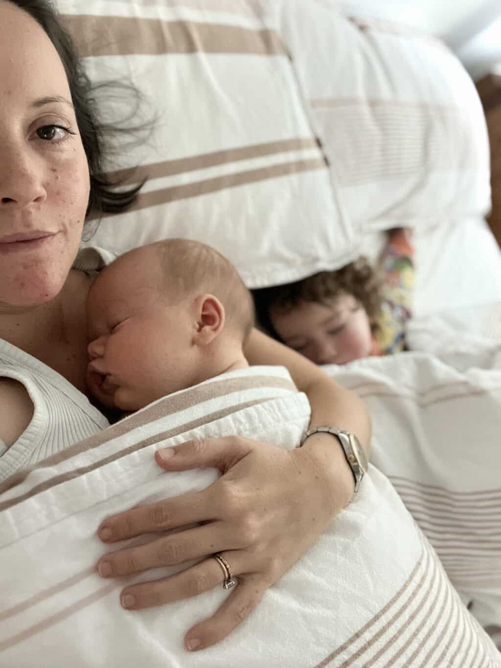 Village For Mama A woman lies in bed holding a sleeping baby while an older child sleeps next to them, partially covered by a blanket.