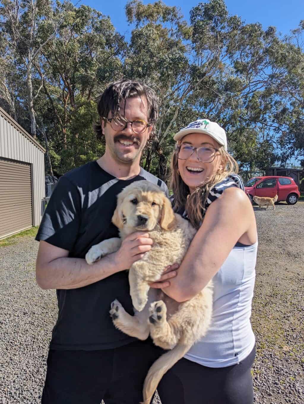 Village For Mama Two people smiling, holding a fluffy puppy outdoors. Trees and a car are visible in the background.