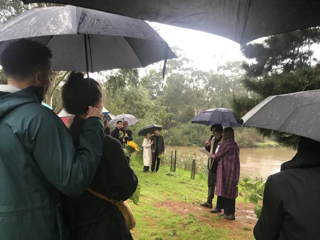 Village For Mama Groups of people holding umbrellas gather outdoors under rain beside a river, listening to a speaker. Some carry flowers and wear raincoats.