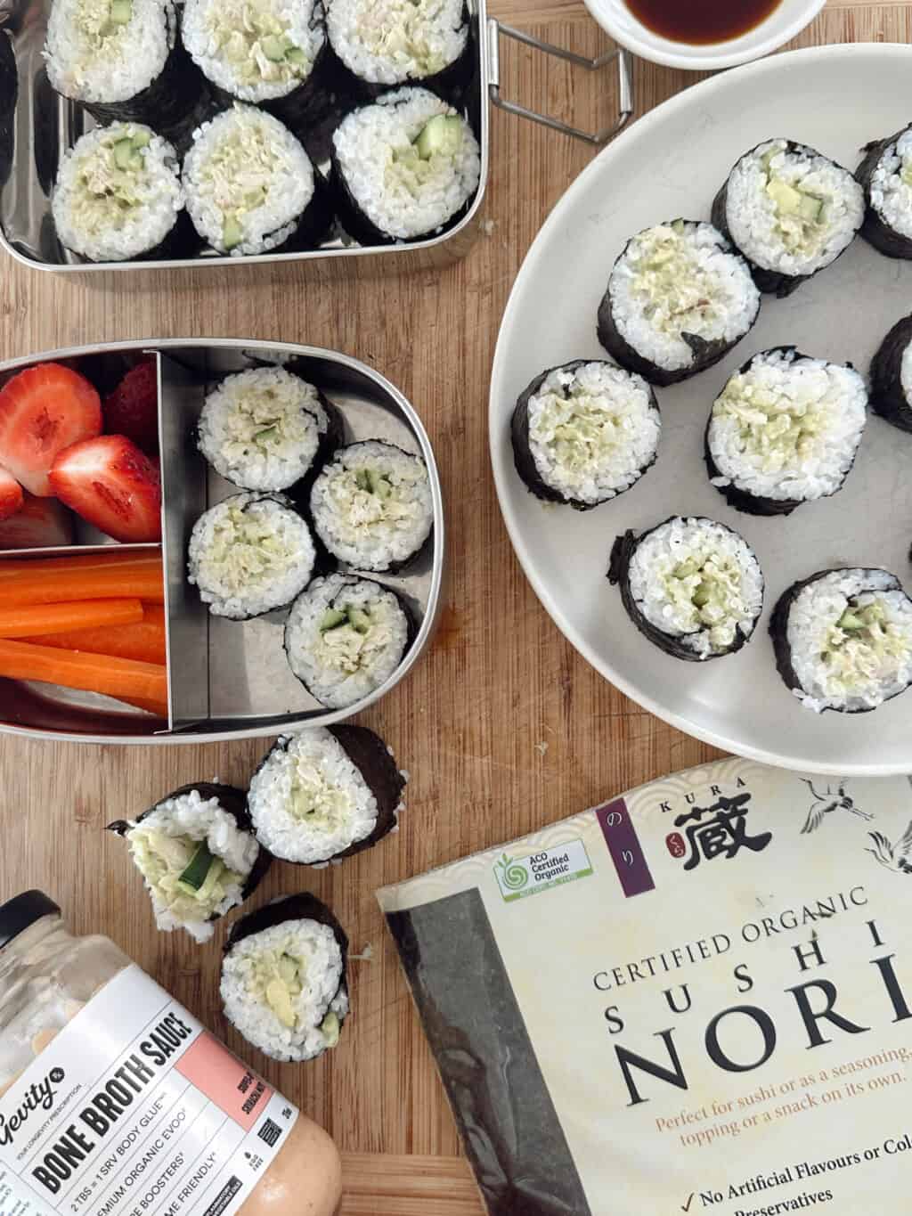 Village For Mama Sushi rolls on a plate and in a bento box with carrots and strawberries. A jar of Bone Broth Sauce and a package of certified organic sushi nori are on the wooden surface.
