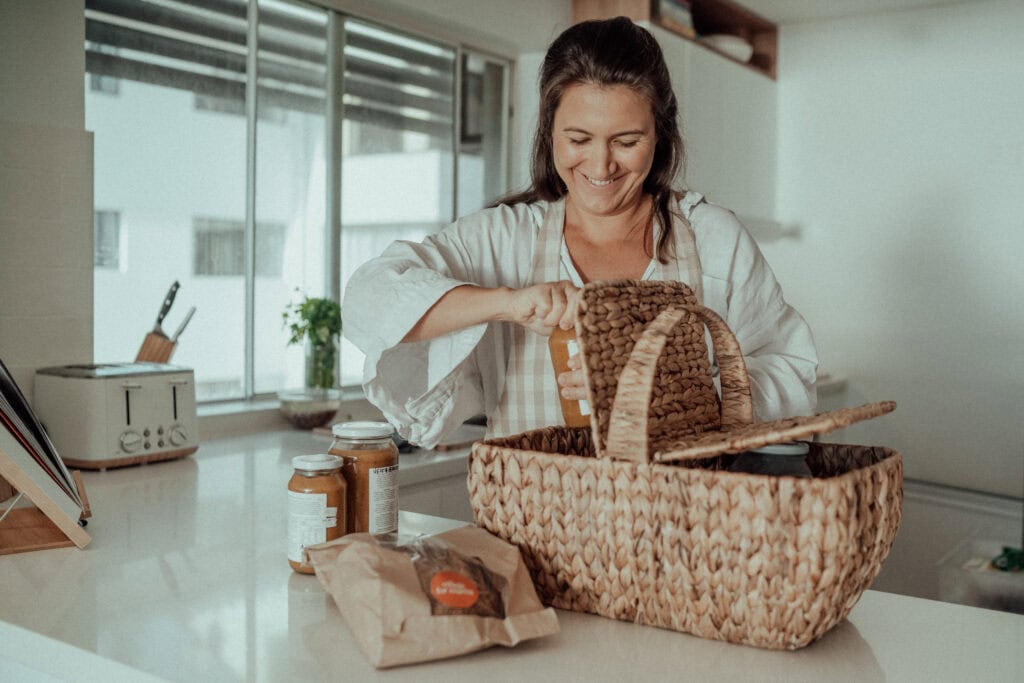 Village For Mama Woman smiling while packing food items into a woven basket on a kitchen counter.