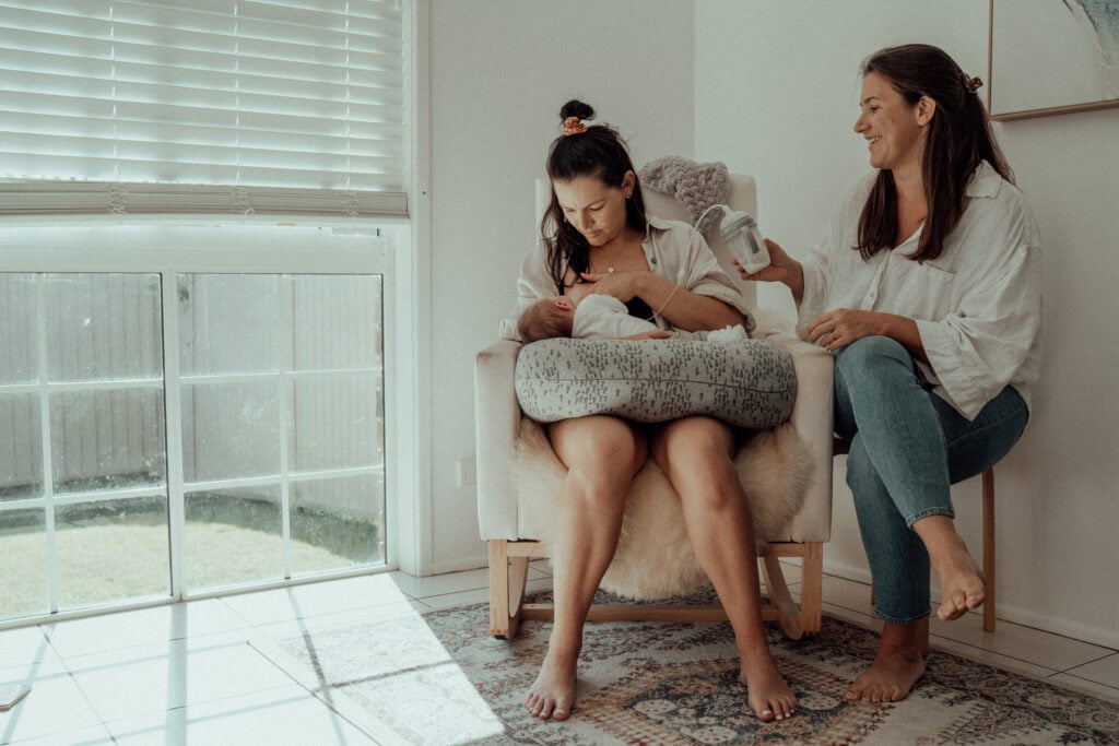 Village For Mama Two women in a sunlit room, one seated and breastfeeding a baby cushion, the other smiling and sitting nearby holding a mug.