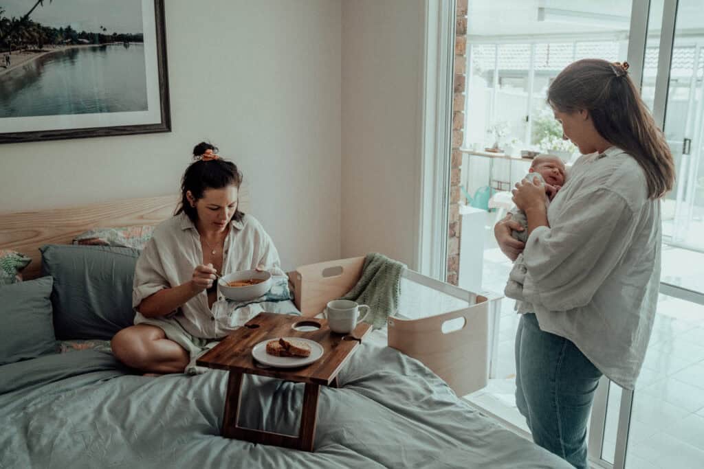 Village For Mama Two women in a bedroom; one is eating on a bed with a tray, and the other is standing, holding a baby. A bassinet is nearby, and a large window lets in natural light.