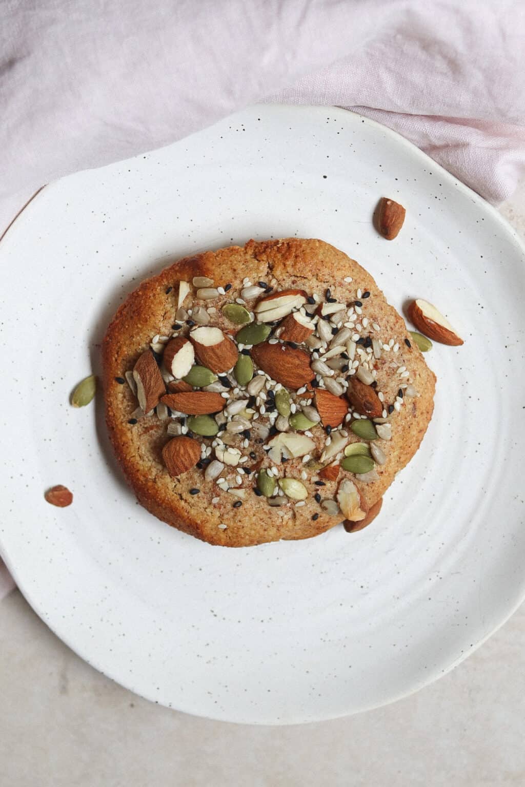 Village For Mama Round cookie topped with assorted nuts and seeds on a white plate.