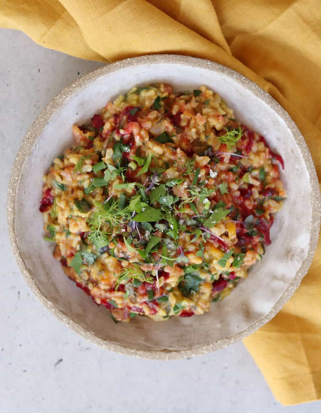 Village For Mama A bowl of colorful vegetable risotto garnished with fresh herbs, placed on a concrete surface next to a yellow cloth.