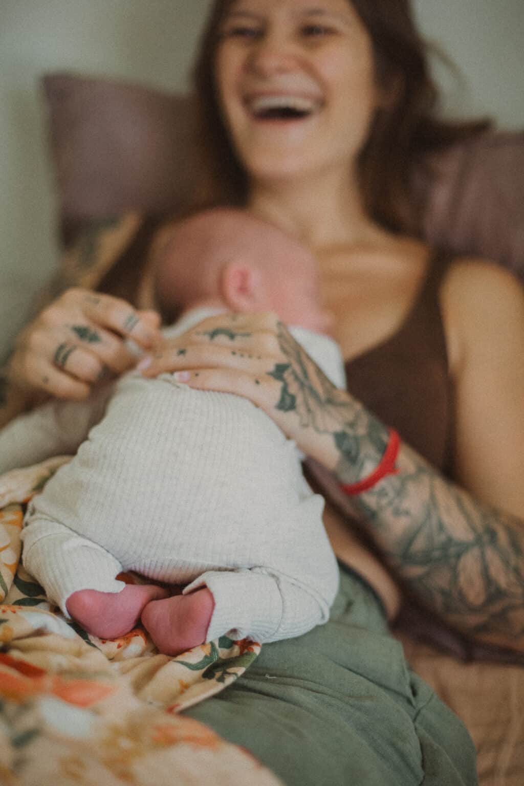 Village For Mama A woman with tattoos is lying down, smiling, and holding a sleeping baby dressed in white.
