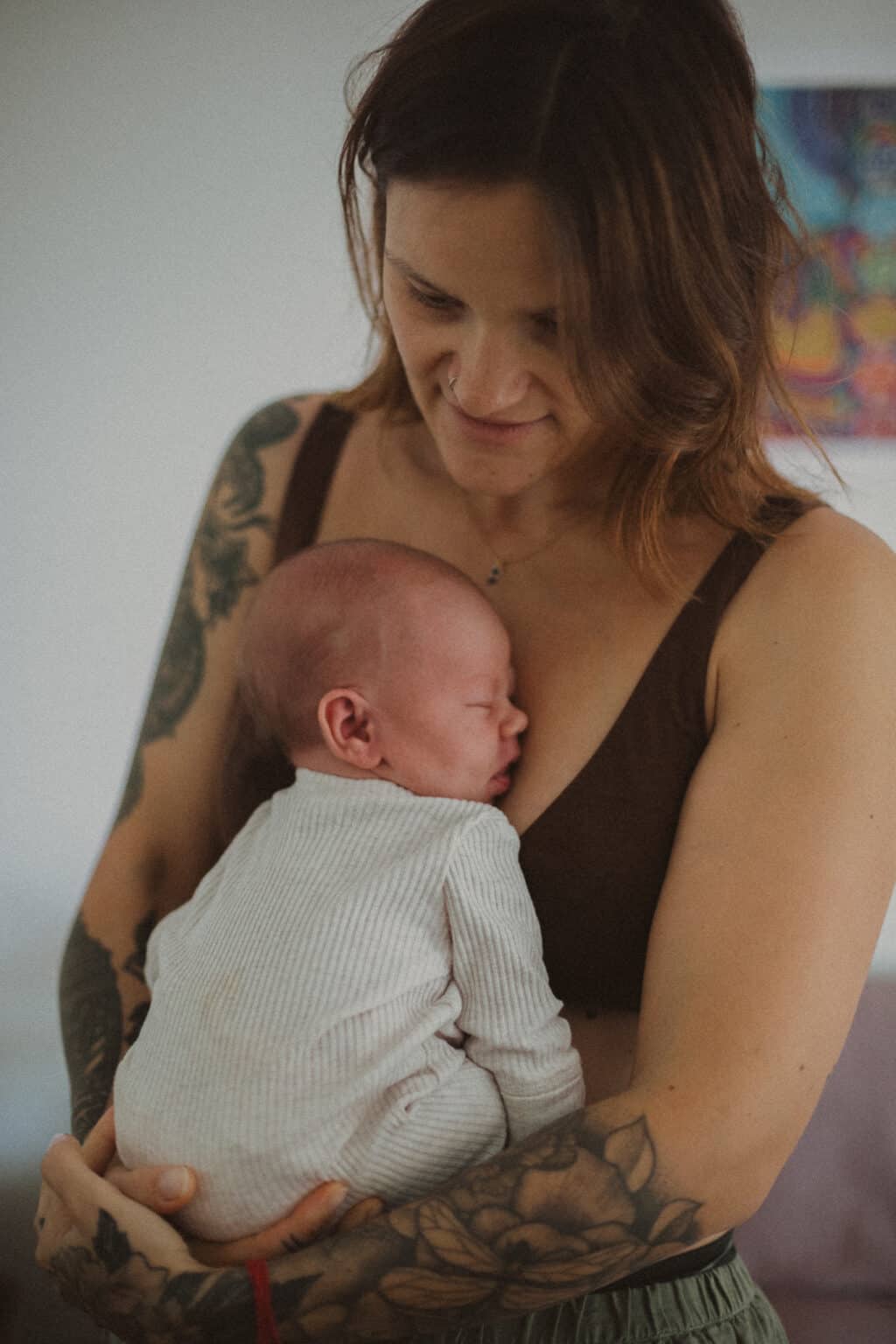 Village For Mama A person with tattoos holds a sleeping baby against their chest in a warm, indoor setting.