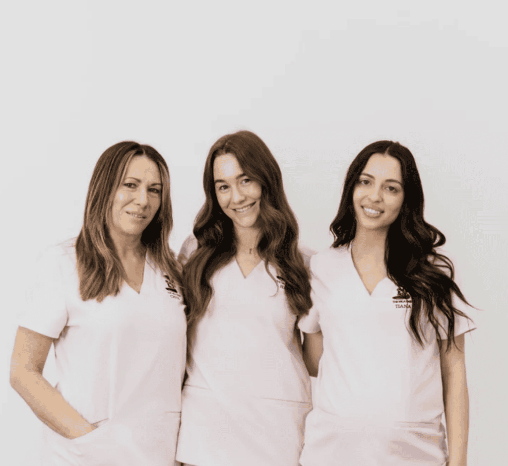 Village For Mama Three women wearing light pink scrubs stand together smiling against a plain white background.