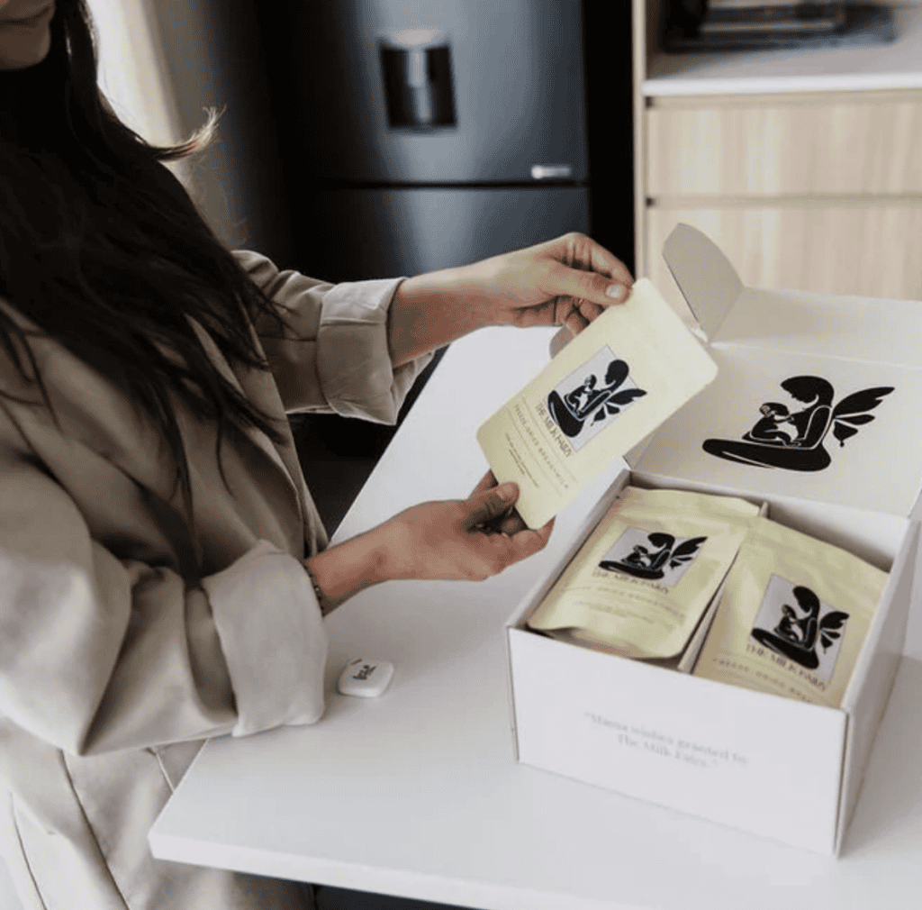 Village For Mama A woman opens a box containing several small packages with stylized illustrations. The packages are light-colored, and the box is placed on a white surface in a kitchen setting.